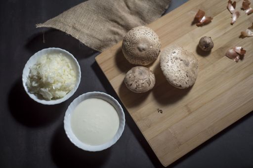 A top view of fresh white mushrooms on a wooden cutting board with chopped onions and sauce in bowls