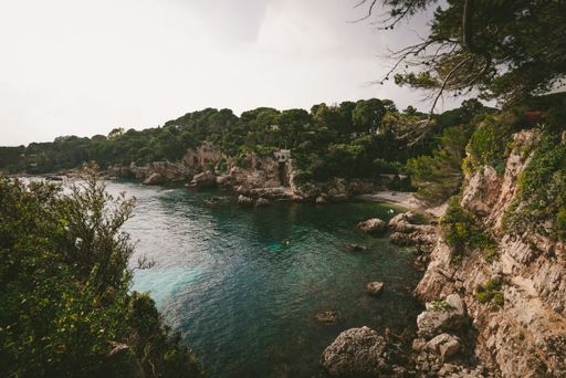 A sea surrounded by rocks covered in greenery under a cloudy sky and sunlight