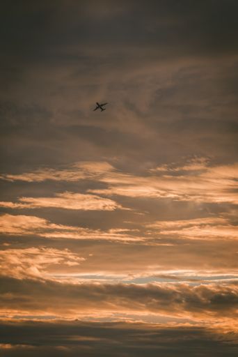 A vertical low angle shot of the airplane flying high in the cloudy sky during the sunset