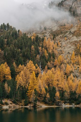 A vertical shot of a calm reflective lake and low flying clouds covering a rough mountain covered with colorful autumn foliage