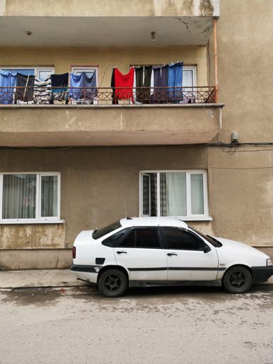 A vertical shot of a white car parked in front of a building with laundry on the balcony