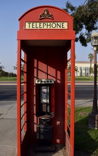 A vertical shot of the front view of an all-red old telephone booth, near a street on a sunny day
