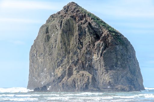 A big rock covered in greenery and fog in a sea under a blue cloudy sky