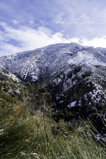 A landscape of mountains surrounded by greenery covered in the snow under a blue cloudy sky