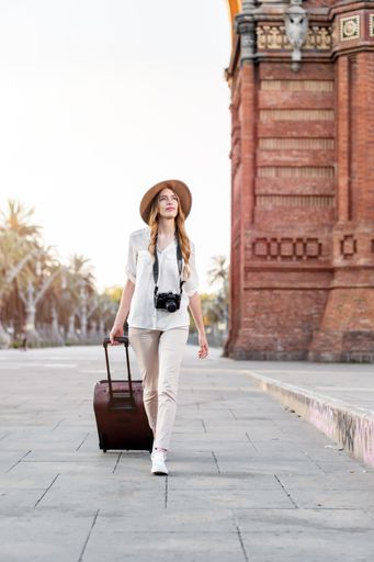 A shallow focus of a female tourist with a suitcase, camera, and a fedora hat walking down Barcelona streets