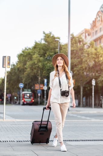 A shallow focus of a female tourist with a suitcase, camera, and a fedora hat walking down Barcelona streets