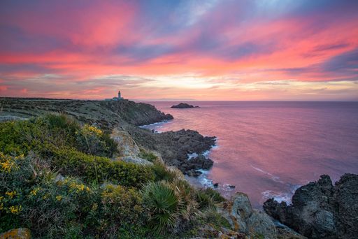 A beautiful scene of a sea with a rocky beach and a lighthouse during a pink sunset