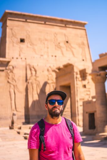 A vertical shot of a Caucasian man wearing a pink t-shirt and posing in front of Philae Aswan, Egypt