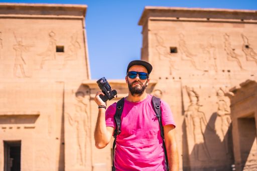 A Caucasian man holding a camera, wearing a pink t-shirt and posing in front of Philae Aswan, Egypt
