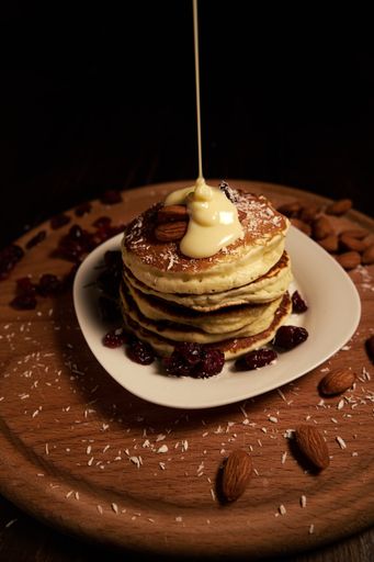A vertical high angle shot of a stack of pancakes with raisins and almonds in a white plate with pouring white syrup on a chopping board