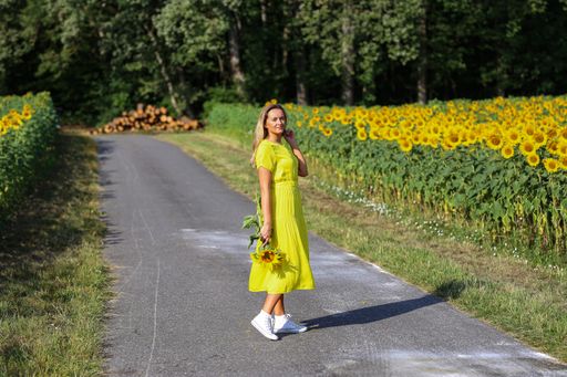 A cute girl in a yellow dress posing with bright sunflowers on the road between the fields of bright flowers
