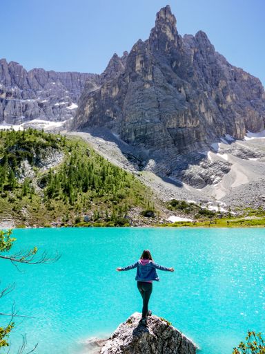 A young Caucasian female standing on a cliff by a lake surrounded by rocky mountains