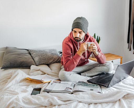 A closeup of a guy sitting on the bed with his laptop and books and a cup of coffee in his hands
