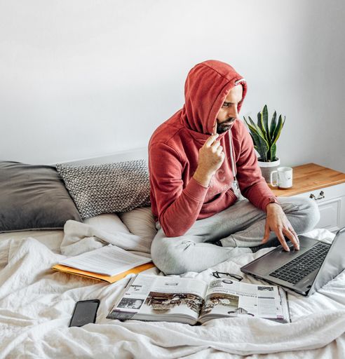 A closeup of a guy in a red hoodie sitting on the bed and working with a laptop