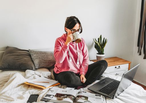 A caucasian female drinking coffee while working with her laptop on a bed