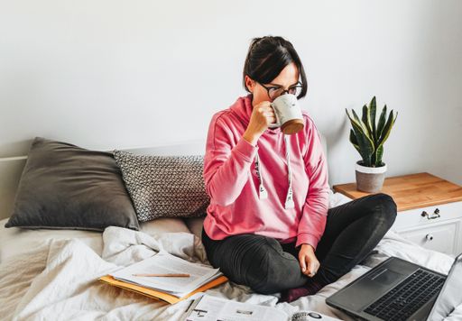 A caucasian female drinking coffee while doing research with a computer and books sitting on the bed at home