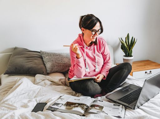 A female doing academic research with a computer and books while sitting on the bed at home