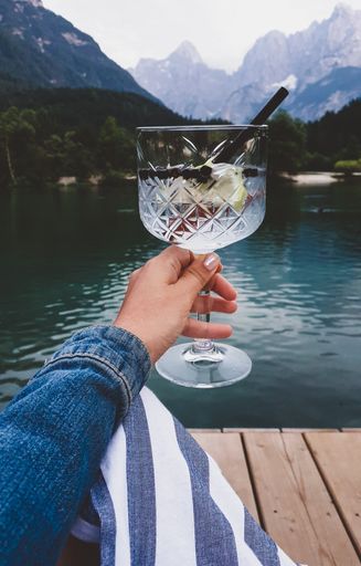 A vertical shot of a female holding a glass of cocktail against a mountainous lake scenery