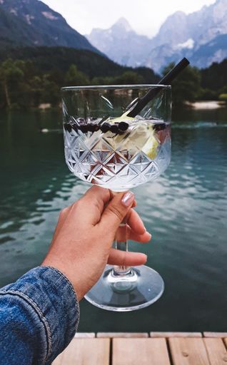 A vertical shot of a female holding a glass of cocktail against a mountainous lake scenery