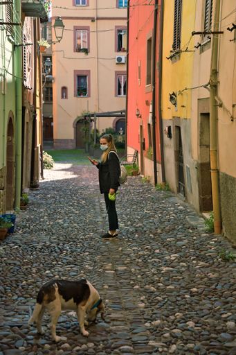 A woman wearing a facemask looking at a dog along a narrow street