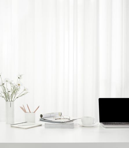 A vertical shot of a cup of coffee, a laptop,  a vase full of flowers, magazines on a white table