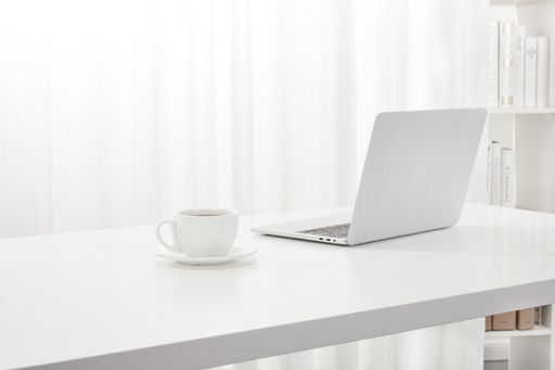 A closeup of a laptop and a cup of coffee on a white desk, next to a bookcase indoors