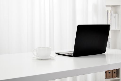 A closeup of a laptop and a cup of coffee on a white desk, next to a bookcase indoors