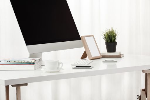 A closeup of a computer and keyboard, a cup of coffee, a smartphone, magazines, and more on a white desk, indoors
