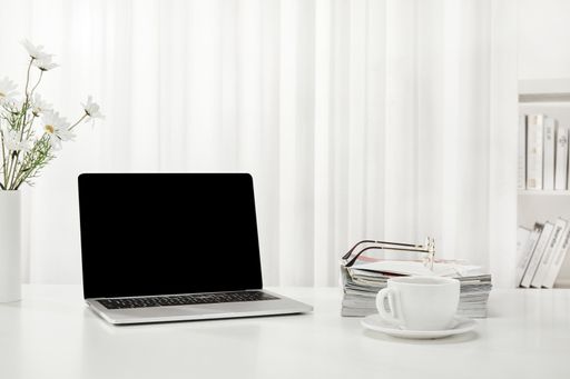 A closeup of a laptop, a cup of coffee, books, magazines, and more on a white desk, indoors