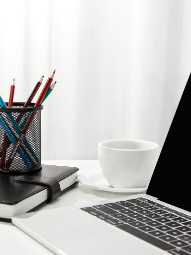 A vertical shot of a laptop, a cup of coffee, a notebook and pencil holder on a white desk, indoors