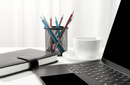 A closeup of a laptop, a cup of coffee, a notebook and pencil holder on a white desk, indoors