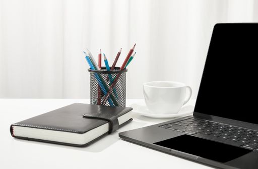 A closeup of a laptop, a cup of coffee, a notebook and pencil holder on a white desk, indoors