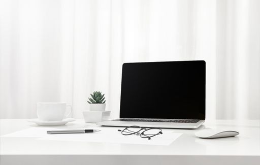 A closeup of a laptop, a cup of coffee, glasses, and more on a white desk, indoors