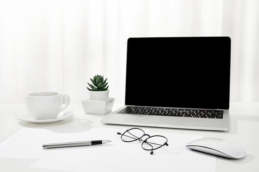 A closeup of a laptop, a cup of coffee, glasses, and more on a white desk, indoors