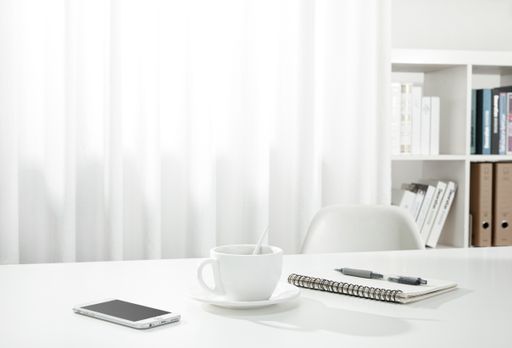 A closeup of a cup of coffee, a notebook and pen, and a smartphone on a white desk, indoors