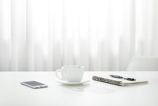 A closeup of a cup of coffee, a notebook and pen, and a smartphone on a white desk, indoors