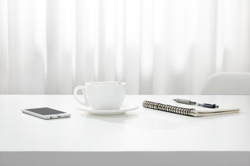 A closeup of a cup of coffee, a notebook and pen, and a smartphone on a white desk, indoors