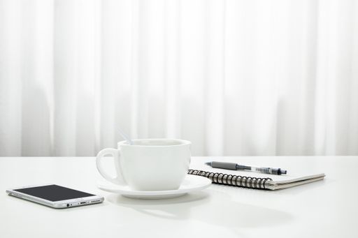 A closeup of a cup of coffee, a notebook and pen, and a smartphone on a white desk, indoors