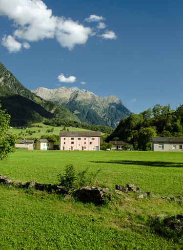 A vertical shot of a field with a white building and scenic rocky mountains in the background