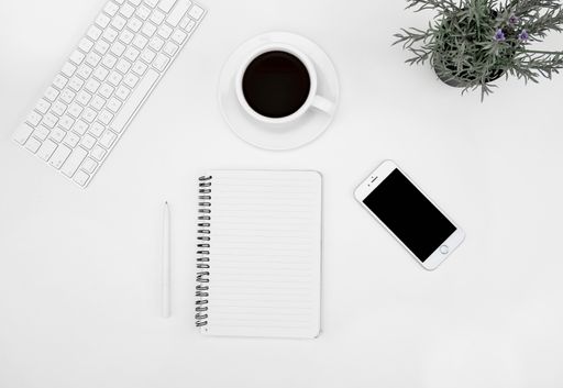 A top view of a cup of coffee, notebook, smartphone, and laptop on a white desk