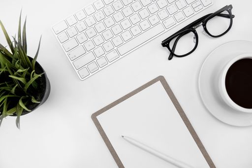 A top view of a cup of coffee, paper and pen, glasses and keyboard on a white desk