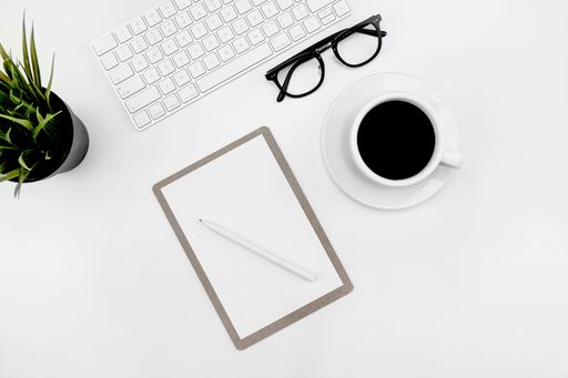 A top view of a cup of coffee, paper and pen, glasses and keyboard on a white desk
