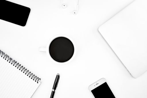 A top view of a cup of coffee, notebook and pen, laptop, smartphone, and more on a white desk