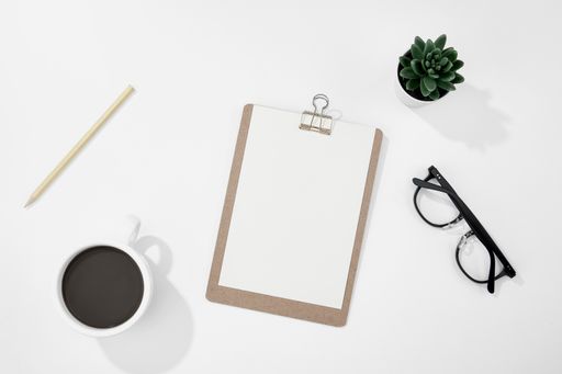 A top view of a cup of coffee, paper and pencil, glasses, and more on a white desk