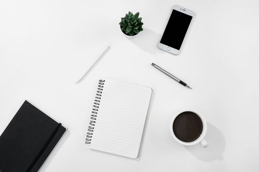 A top view of a cup of coffee, notebook and pen, and a smartphone on a white desk