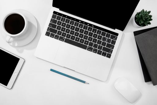 A top view of a cup of coffee, notebook, laptop, and tablet on a white desk
