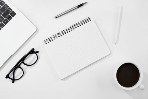 A top view of a cup of coffee, notebook, glasses, and laptop on a white desk
