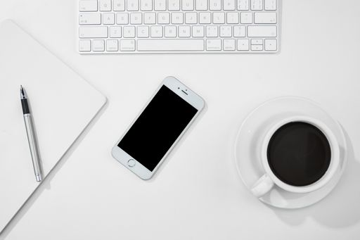 A top view of a cup of coffee, notebook, smartphone, and laptop on a white desk