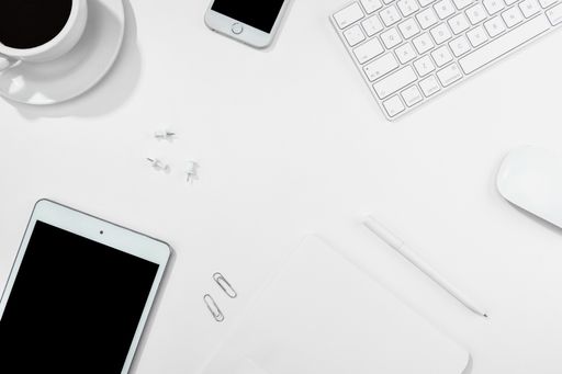 A top view of a cup of coffee, tablet, smartphone, keyboard, and more on a white desk