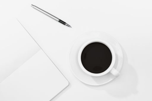 A top view of a cup of coffee, notebook, and pen on a white desk